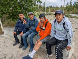Four people sitting together on a bench on sandy grounds outside with trees in background